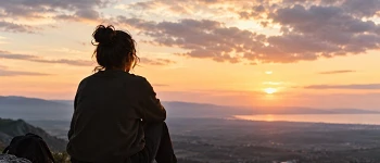 Persona sentada de espaldas sobre unas rocas, contemplando un paisaje montañoso y el mar al atardecer, bajo un cielo cubierto de nubes iluminadas por una luz cálida.